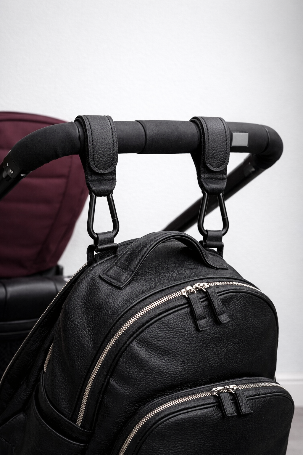 Black leather backpack attached to a stroller handle using a pair of the Mira Buggy clips on a light gray background