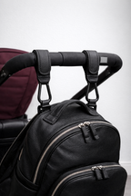Black leather backpack attached to a stroller handle using a pair of the Mira Buggy clips on a light gray background